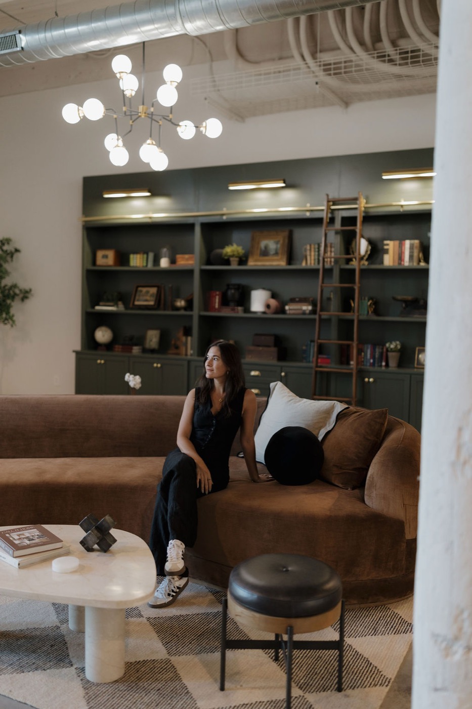 Woman seated on a curved brown velvet sofa in a loft lounge with a globe chandelier and dark green library shelving