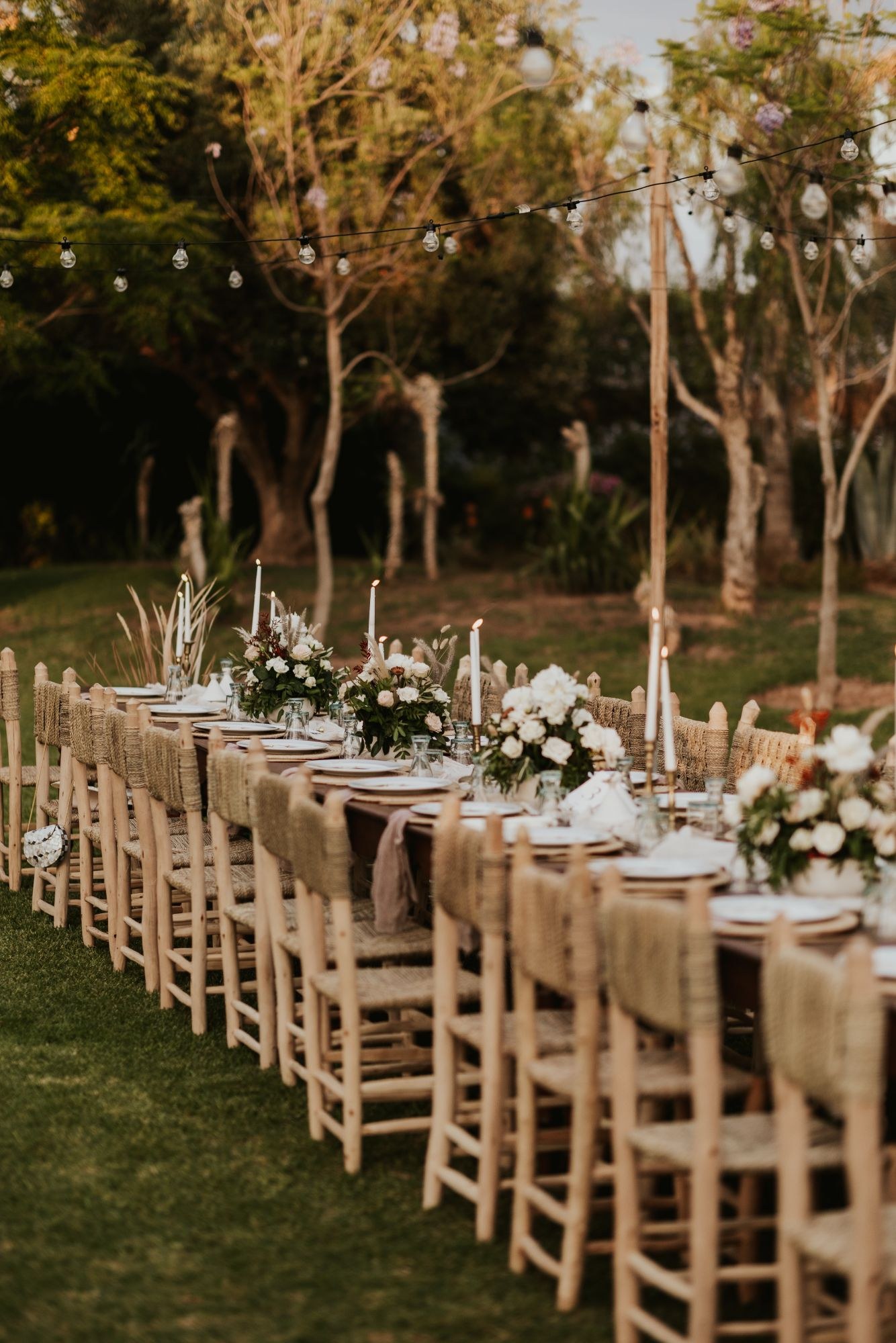 Long candlelit table with woven chairs and white floral arrangements in a garden at dusk, under string lights