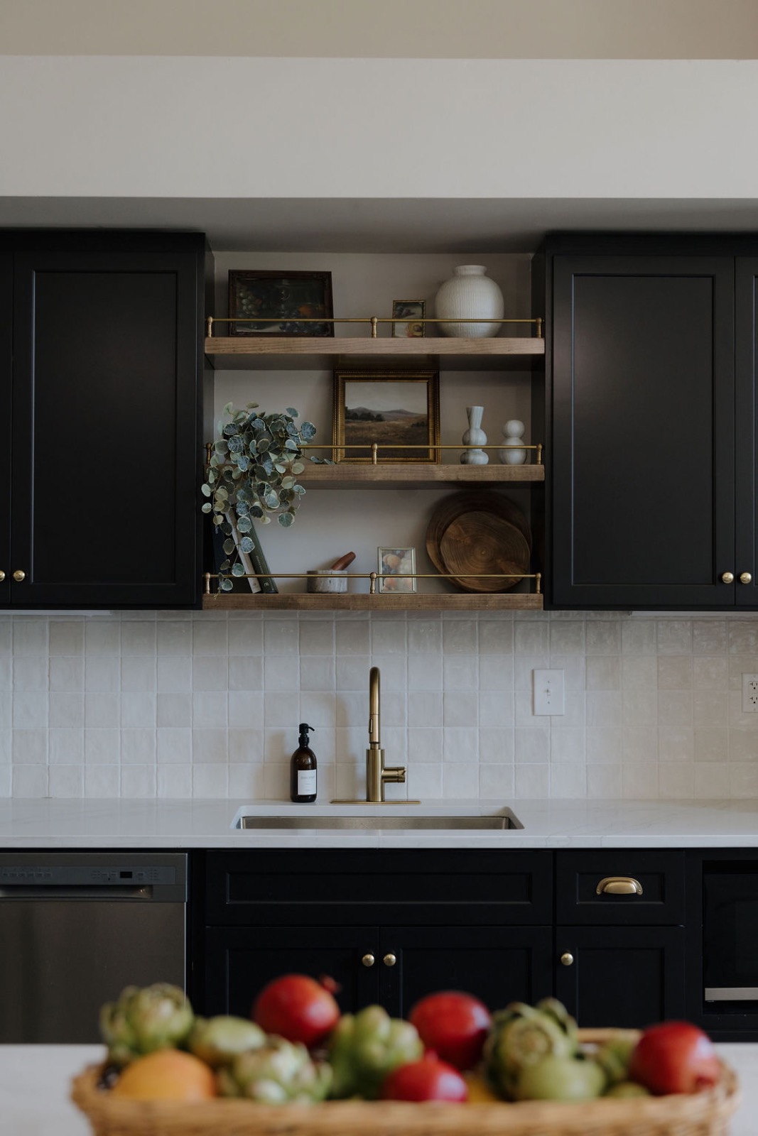 Black shaker cabinets with open wood shelves, brass rails, vintage art, and a brass faucet over a handmade-tile backsplash