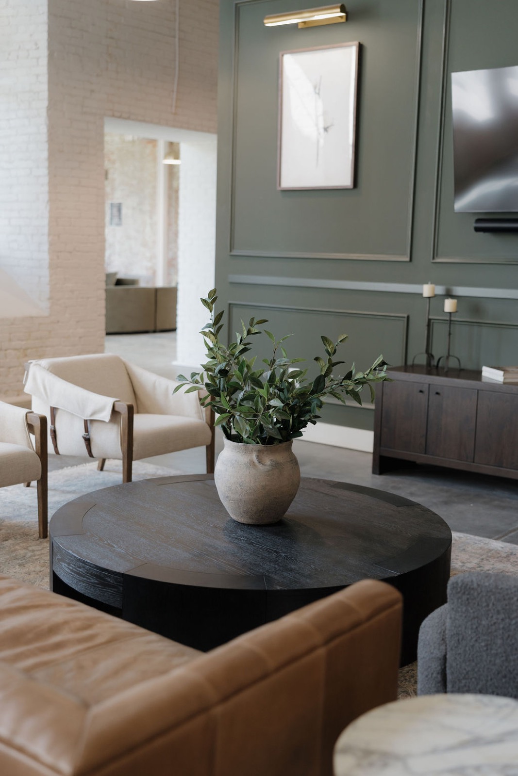 A round black coffee table with an olive-branch vase in front of a green paneled wall and cream armchairs