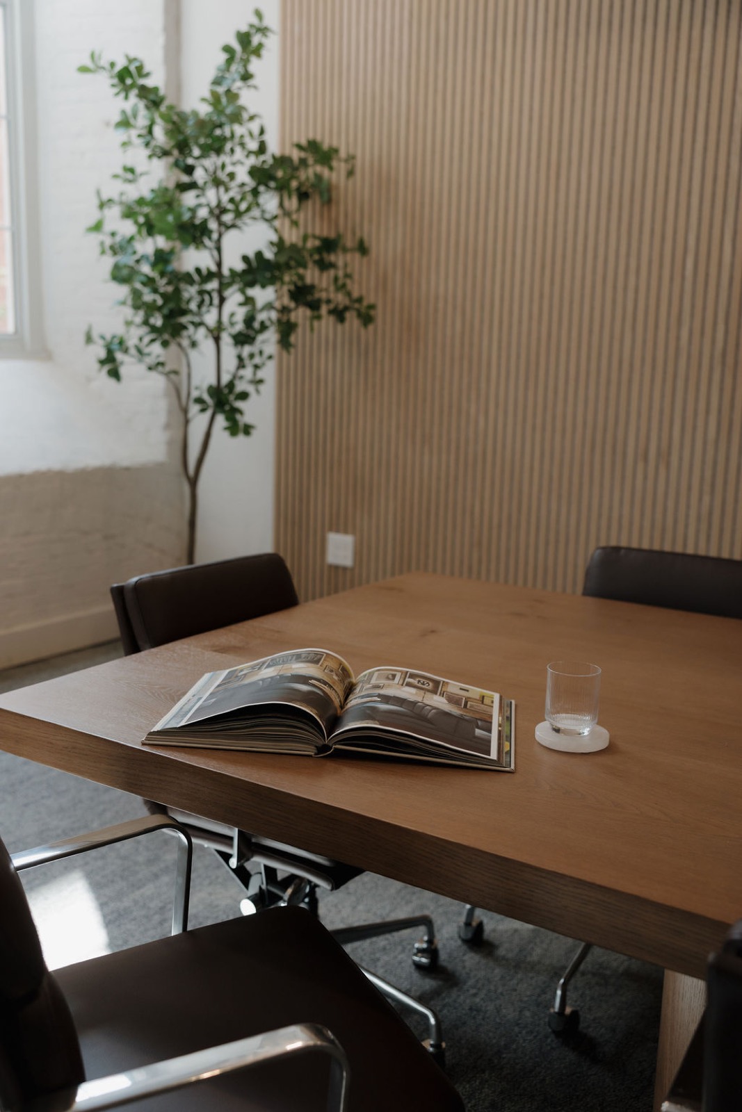 A conference table with an open design book and a glass on a coaster beside a wood slat wall and a tall plant