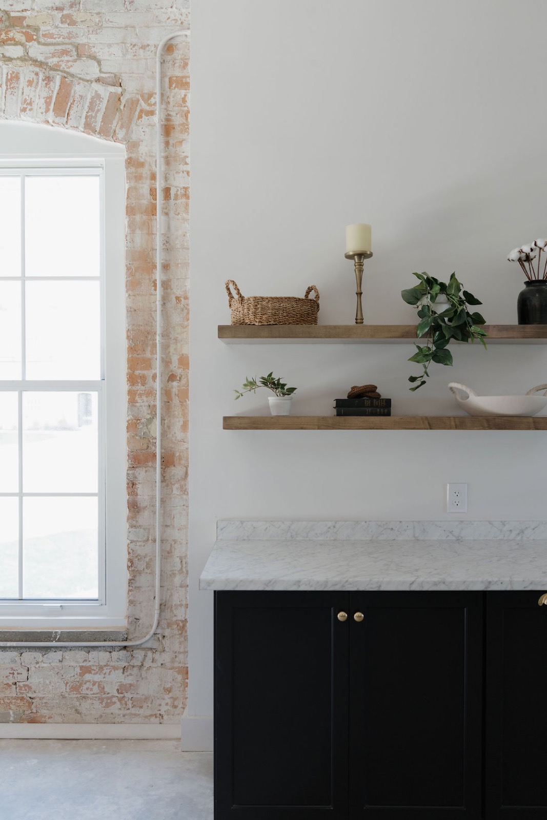 Open wood shelves with a candle, a basket, and plants above a marble counter and black cabinets beside an arched brick window