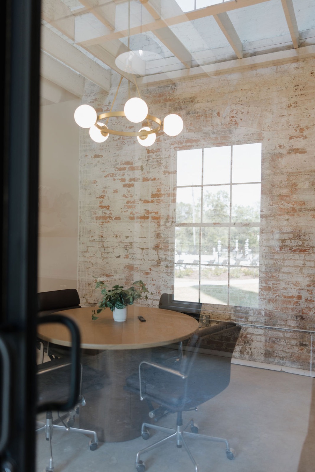 A small meeting room with a round wood table, a brass globe chandelier, and a whitewashed brick wall with a window