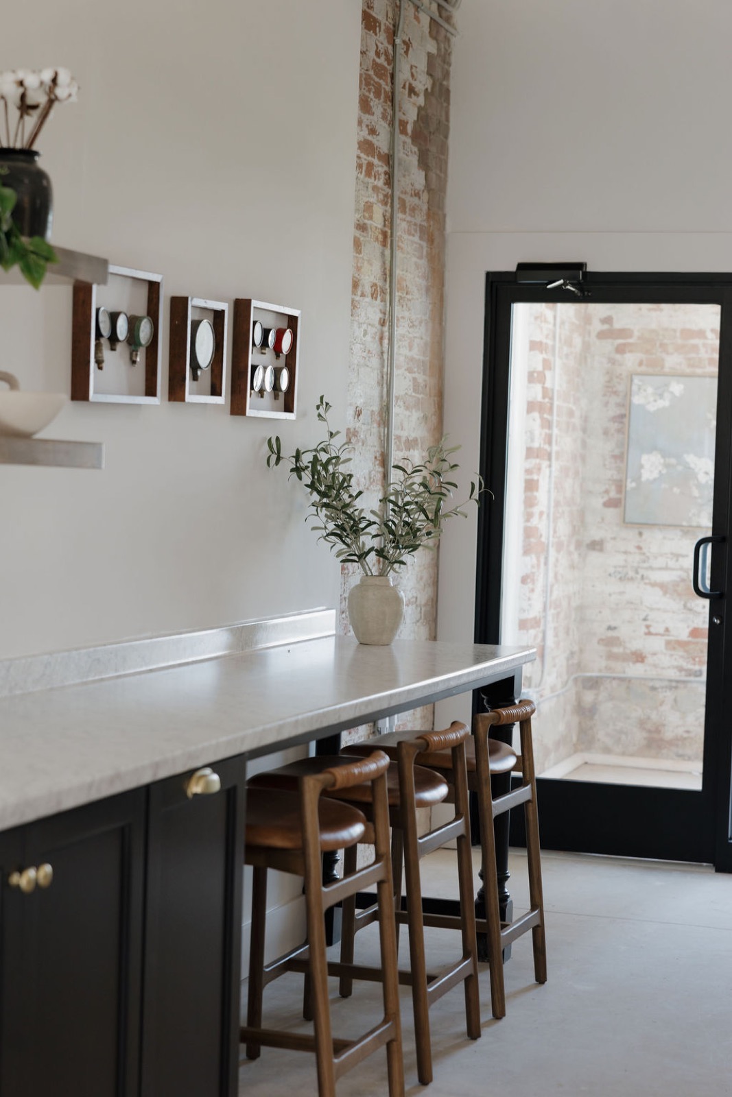 A long marble counter with leather-seat stools, framed vintage gauges, and olive branches beside a black glass door