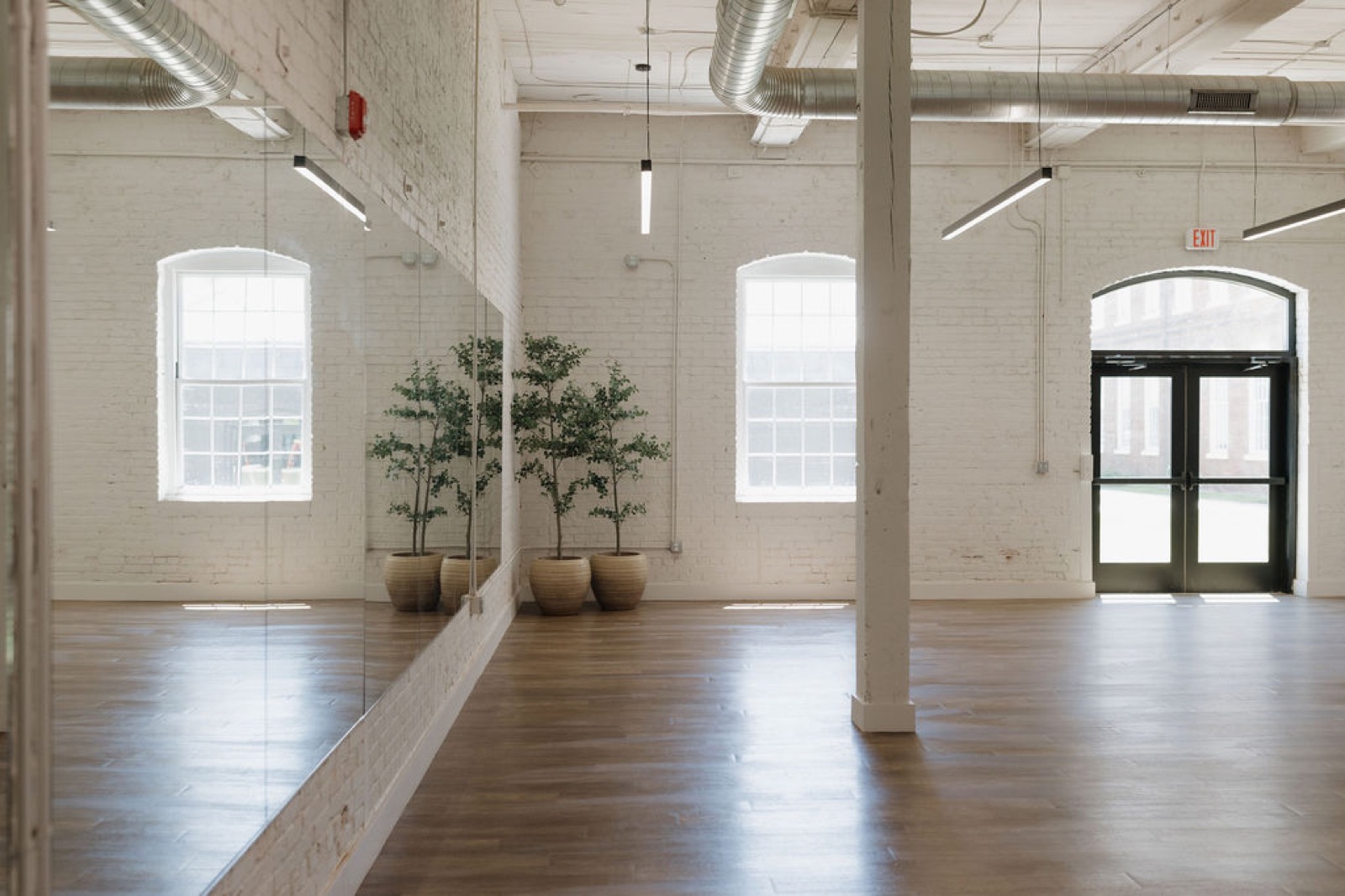 The yoga studio with a mirrored wall, potted trees, arched windows, and a wood floor beneath exposed ductwork