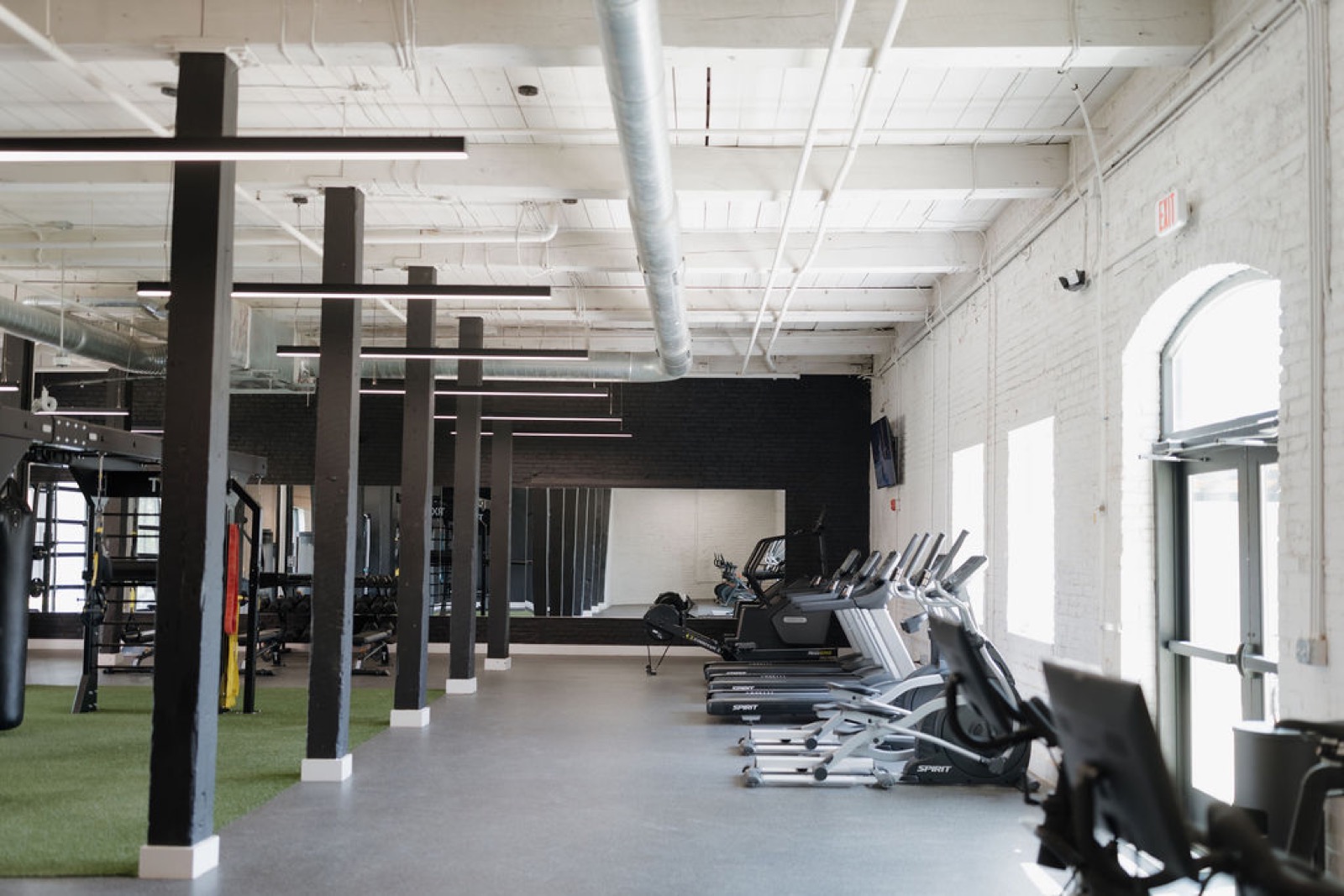 The fitness room with black columns, cardio machines along arched windows, and a turf strip with rigs beyond