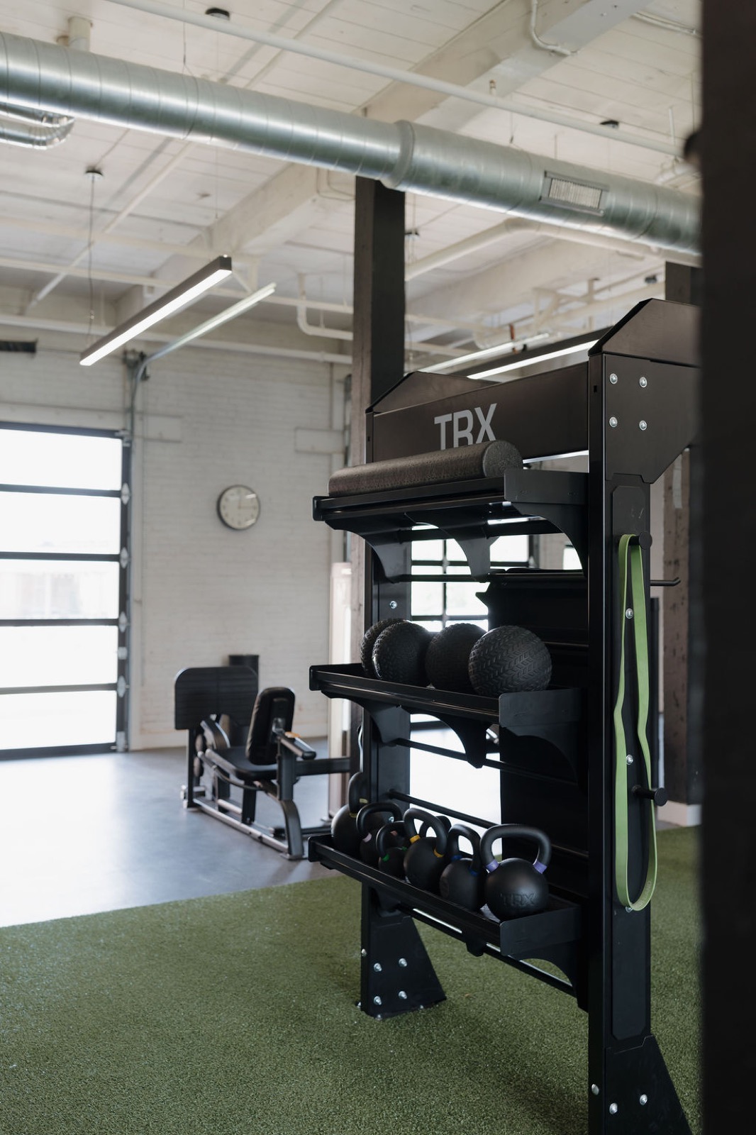 A black storage rack of kettlebells, slam balls, and a foam roller on turf, with a leg press by a glass garage door