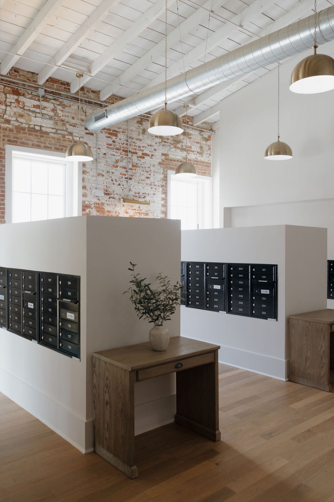 A mailroom with black mailboxes set into white walls, wood console tables, and brass dome pendants beneath exposed brick