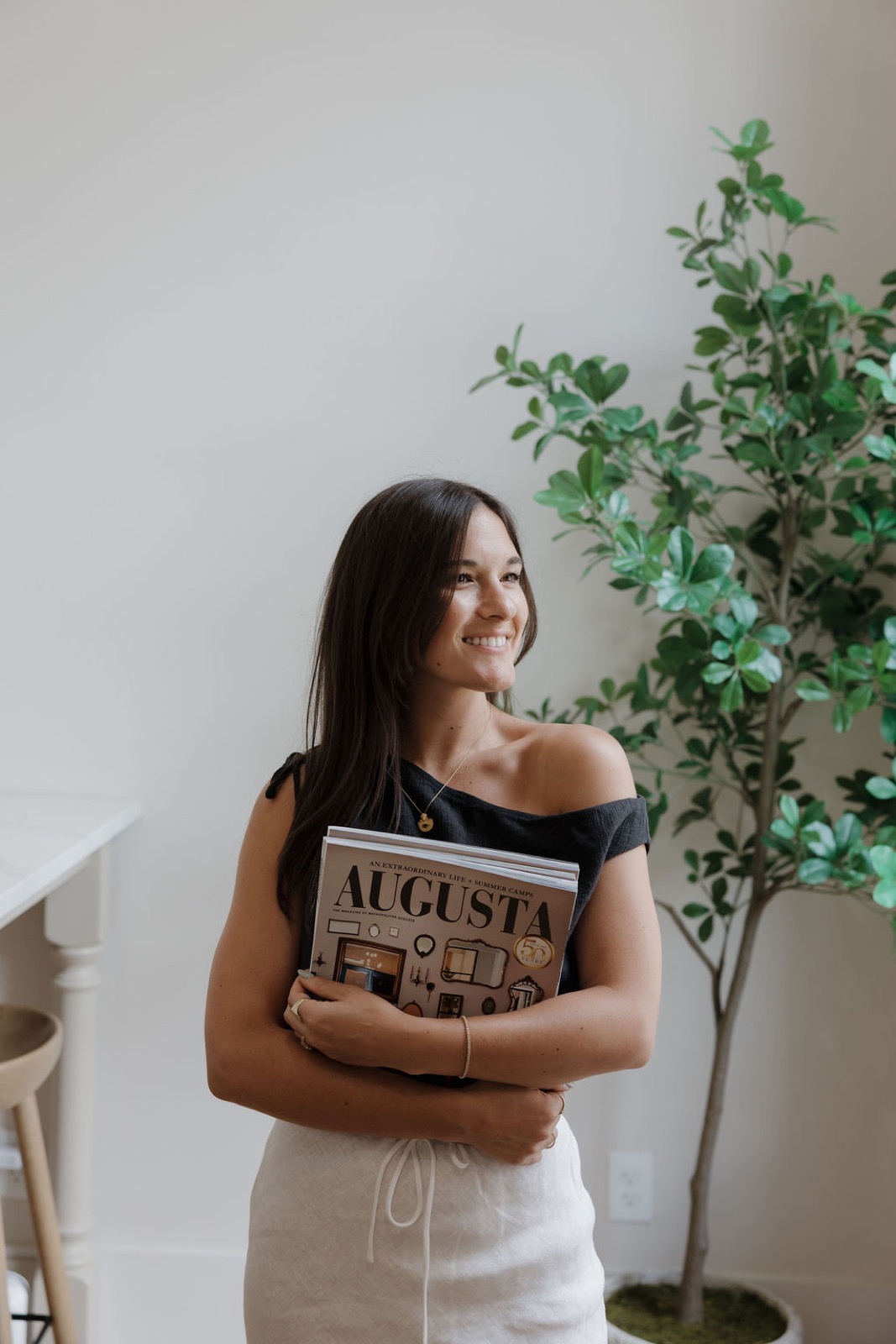 A woman smiling and holding a stack of Augusta magazines beside a potted tree
