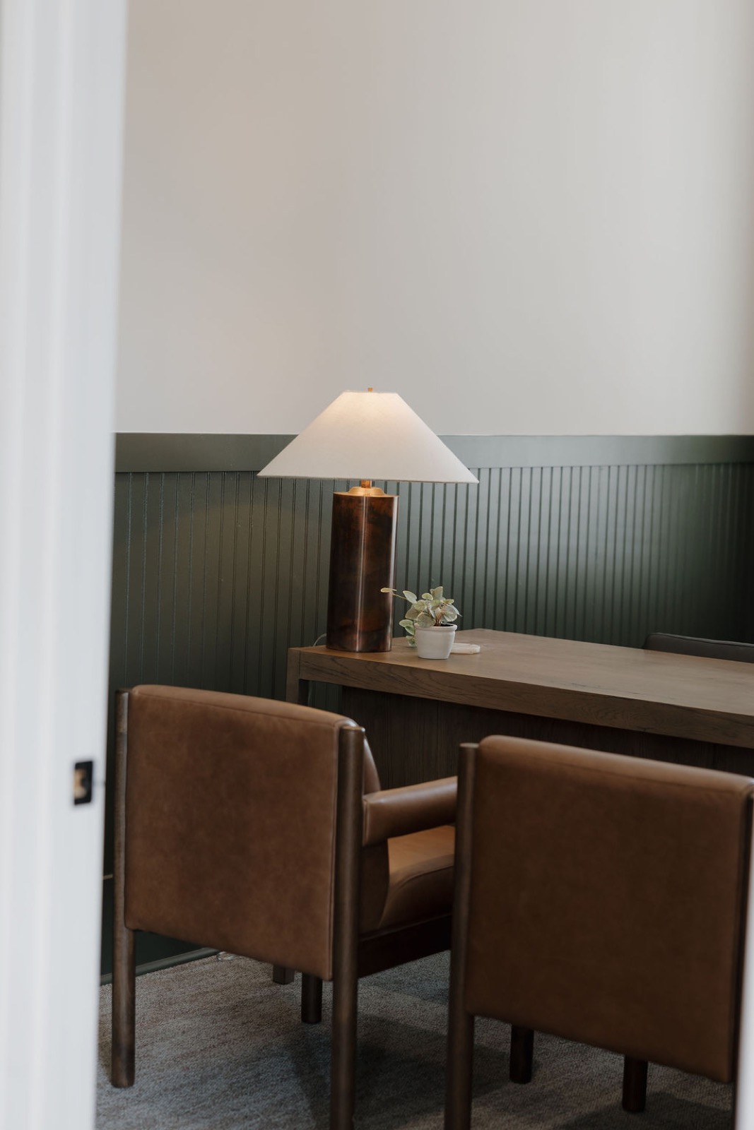 A private office with green beadboard wainscot, a lamp on a wood desk, and tan leather chairs