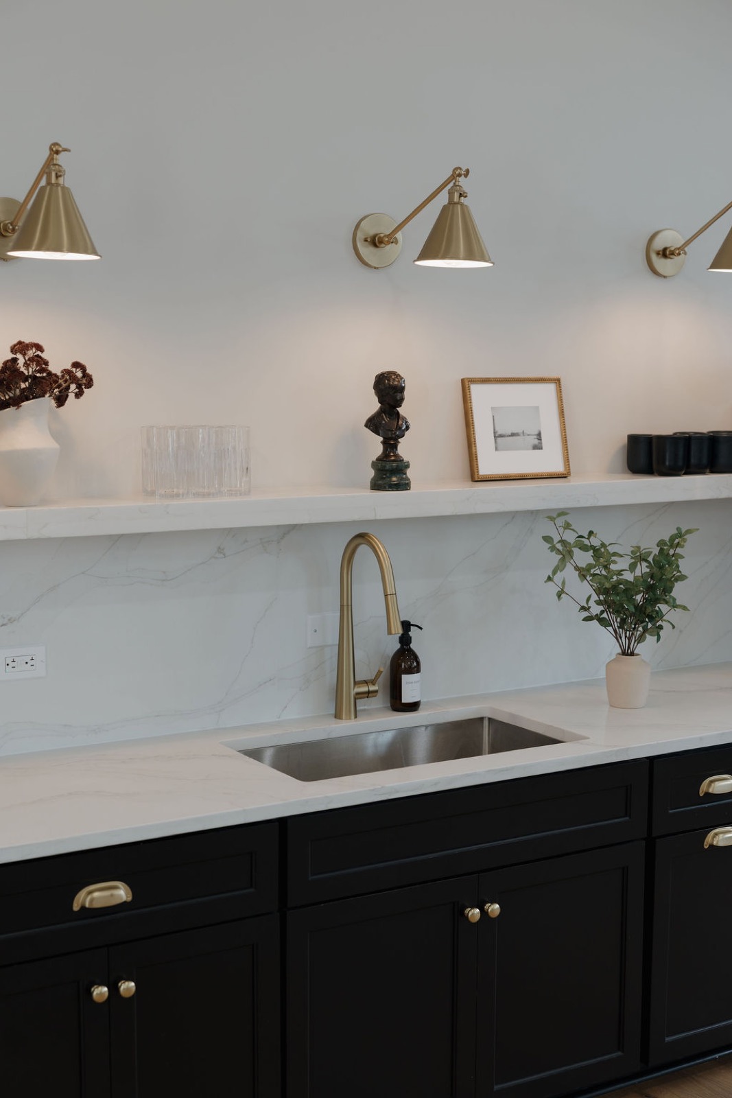 A coffee bar with a marble backsplash, brass wall sconces, a brass faucet, and black cabinets