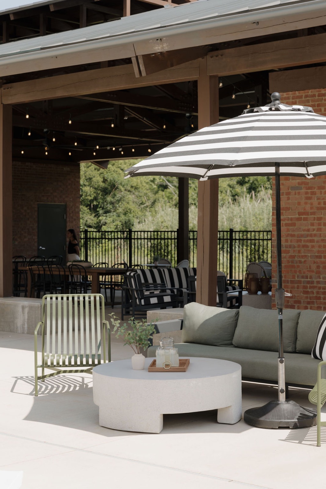 Outdoor lounge with a gray-green sofa, a round white coffee table, olive chairs, and a striped umbrella under a timber pavilion