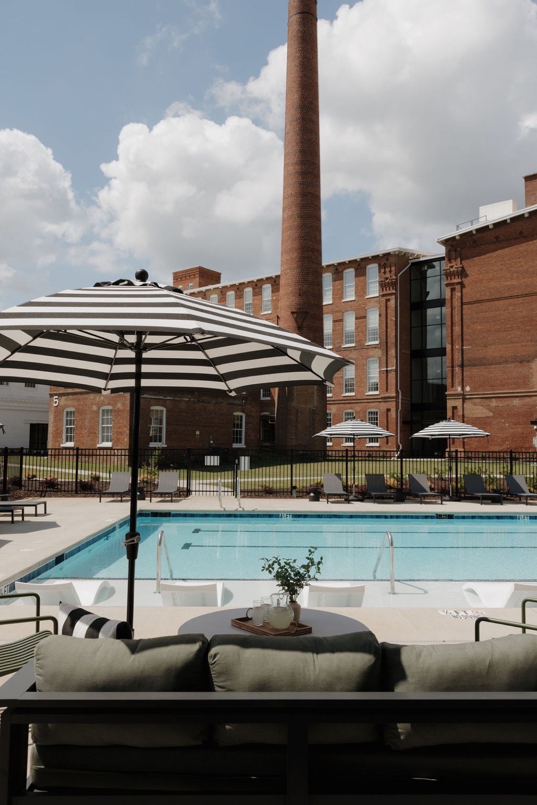 The pool with a striped umbrella and the tall brick smokestack behind a sofa in the foreground