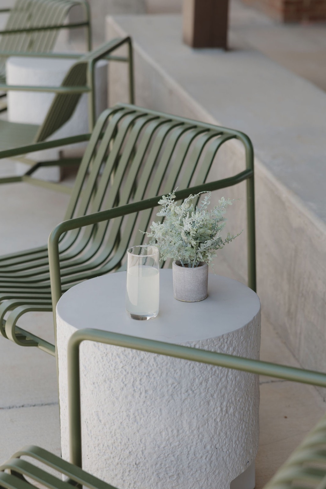 An olive metal lounge chair beside a textured white side table with a glass of lemonade