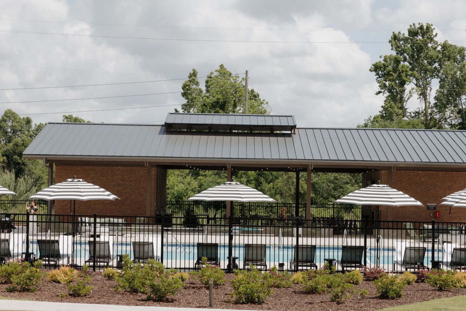 The brick pool pavilion with a metal roof seen across the pool and striped umbrellas