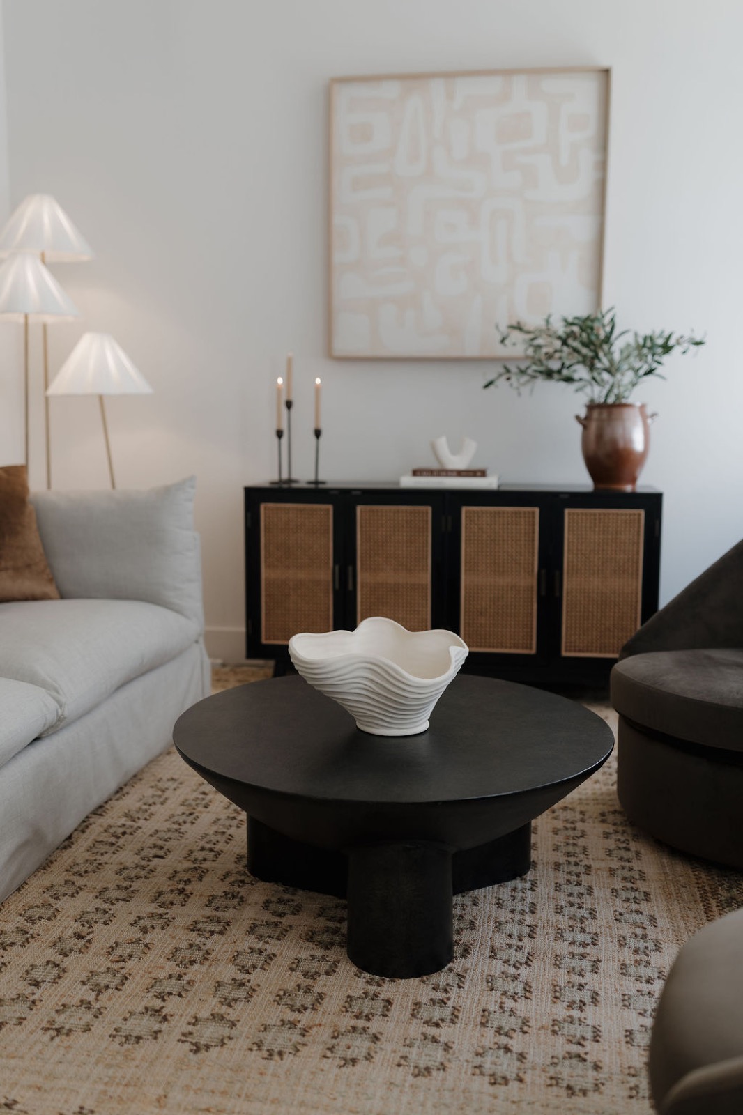 A living room with a gray slipcovered sofa, a black round coffee table with a white wavy bowl, a cane-front sideboard, and abstract art