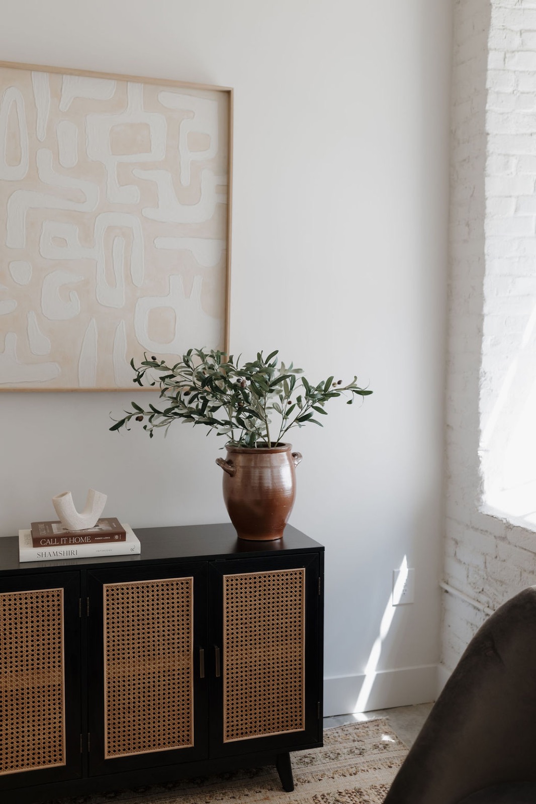 An olive branch in a terracotta crock on a black cane-front sideboard beneath large abstract art, beside a white brick wall
