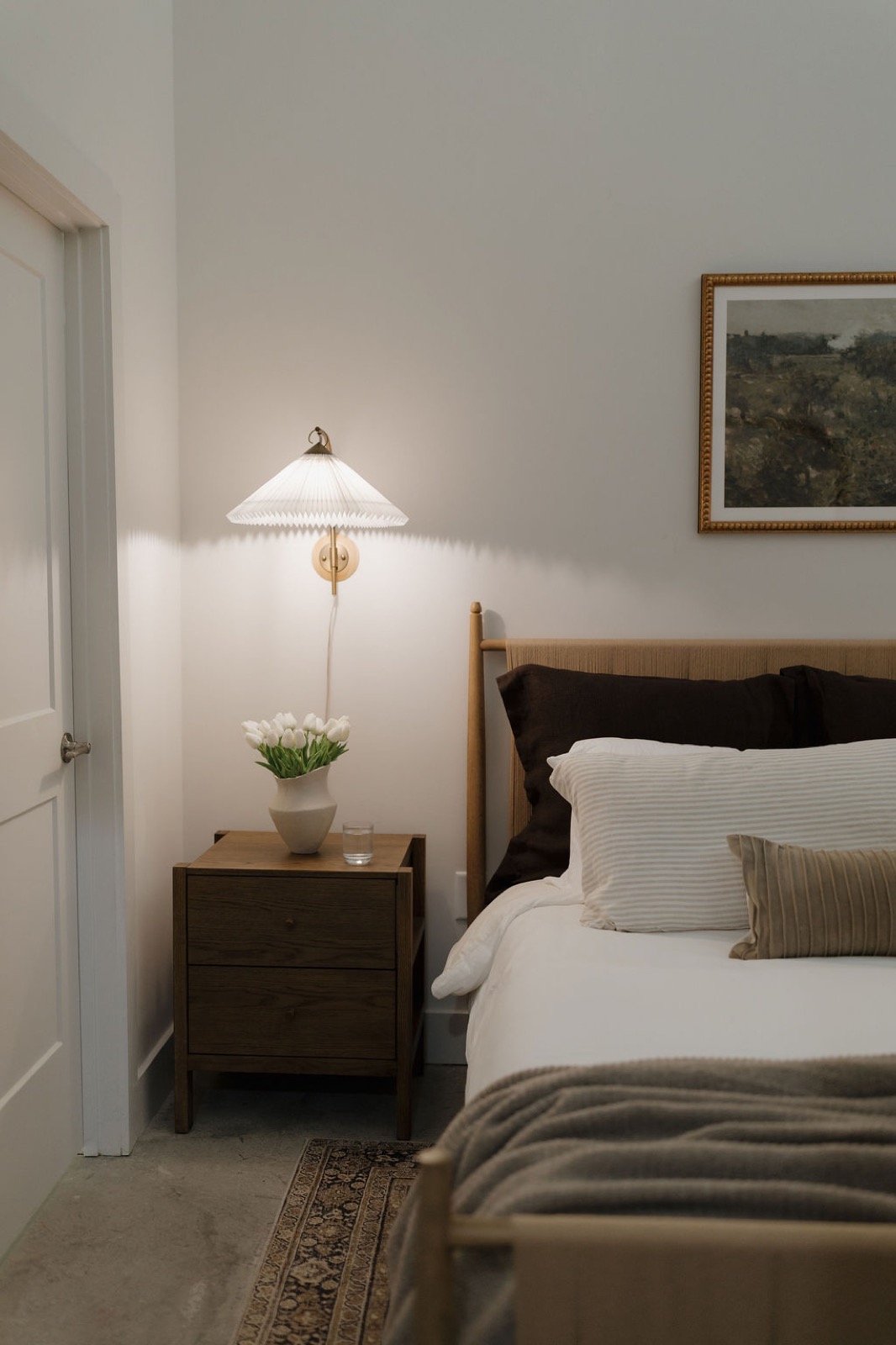 A bedside table with tulips and a pleated wall sconce beside a wood-framed bed with layered neutral bedding