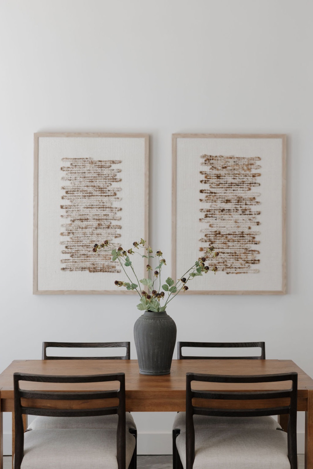 Two woven abstract panels above a wood dining table with black chairs and a gray vase of blackberry branches