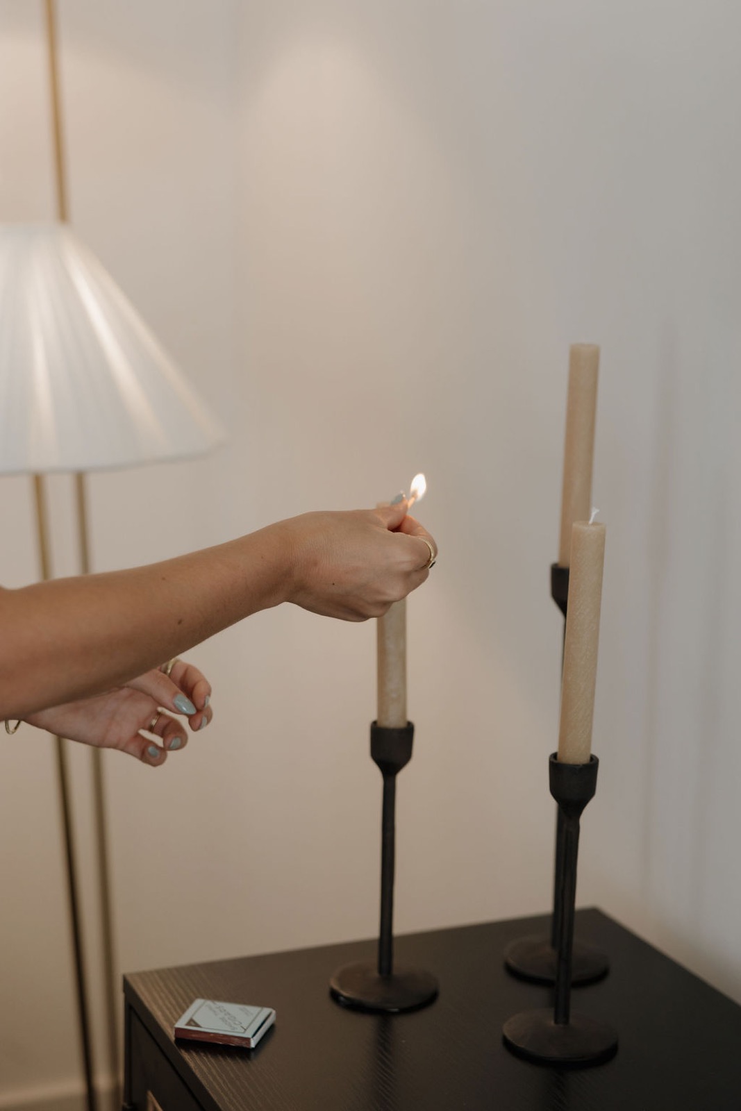 A hand lighting a taper candle on a black console beside a pleated floor lamp