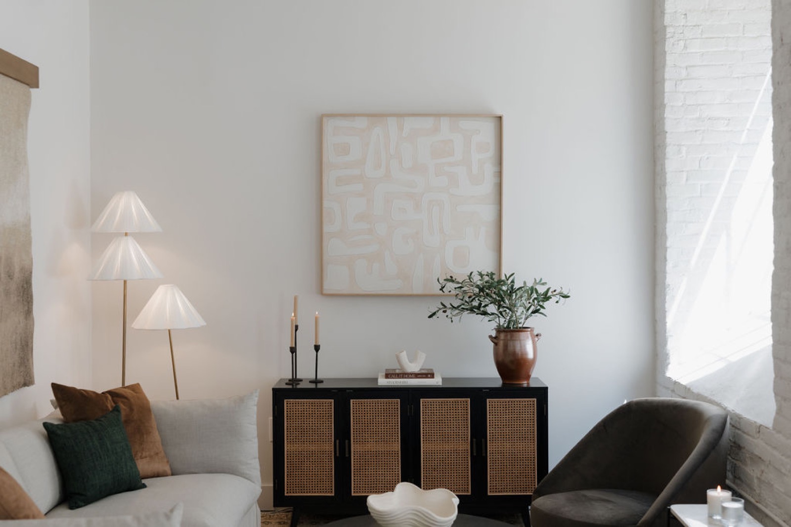 A living room corner with a pleated three-shade floor lamp, a cane-front sideboard, abstract art, and a gray sofa