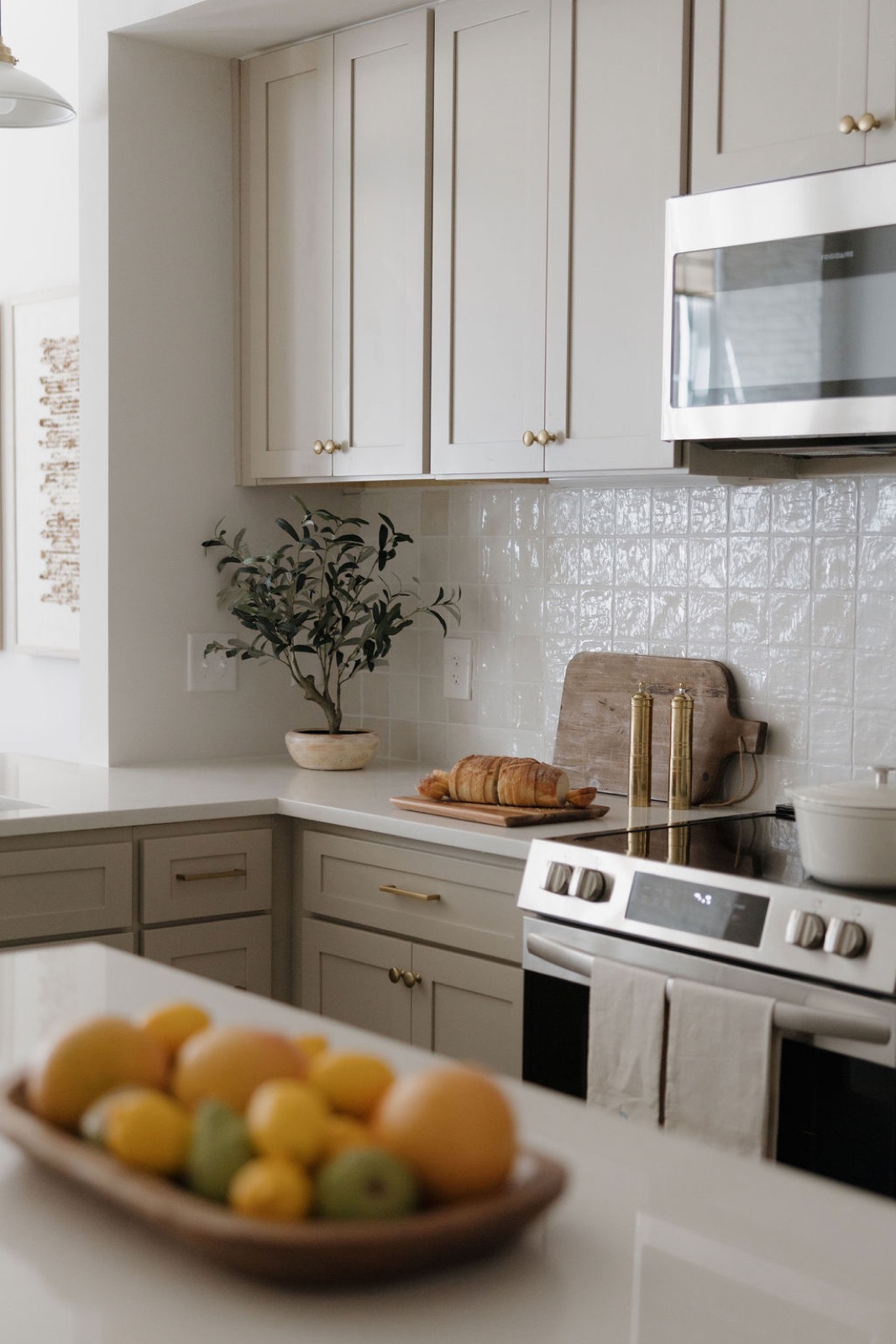 A kitchen corner with greige shaker cabinets, a glazed tile backsplash, brass pepper mills, and a bowl of citrus