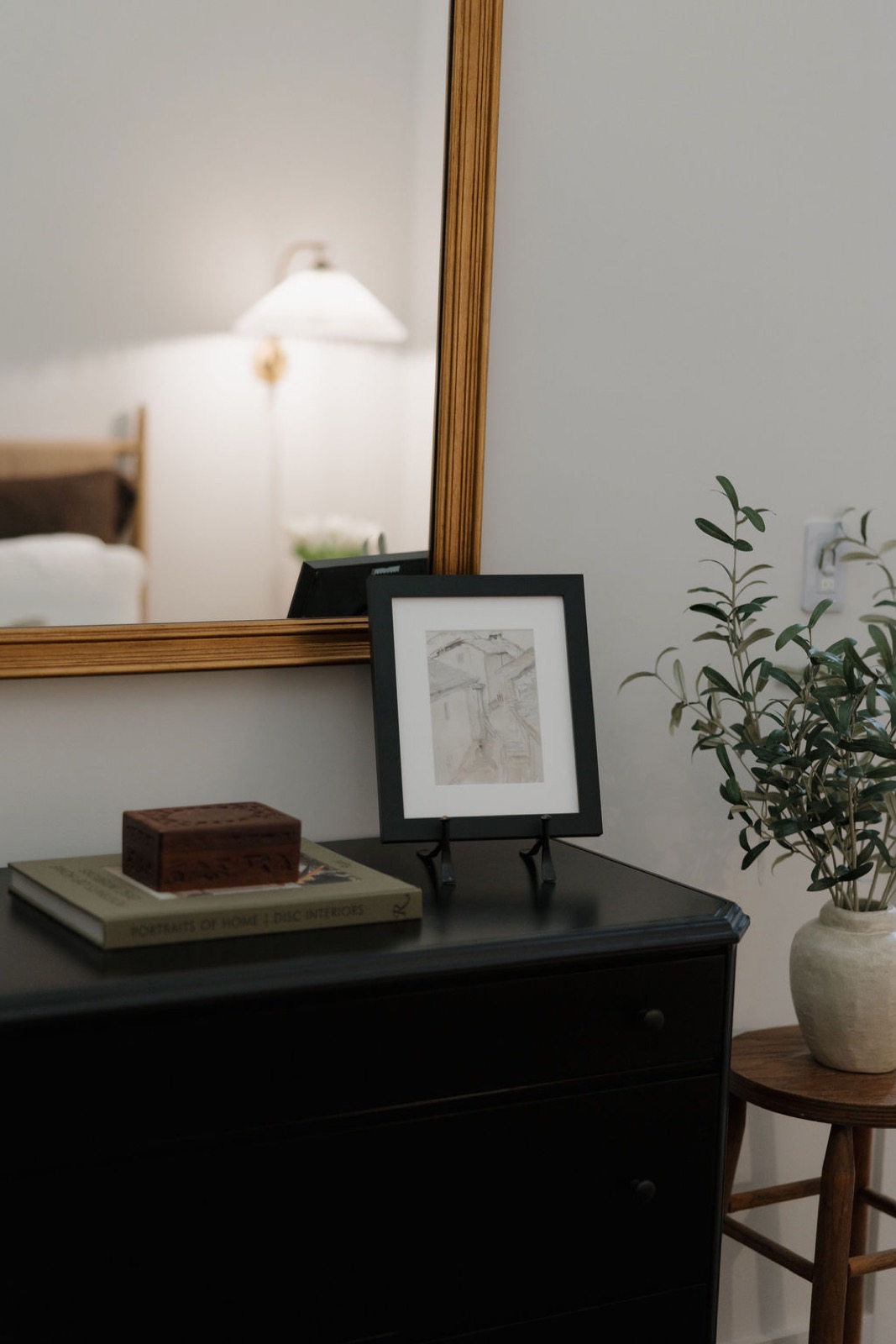 A black dresser with a framed sketch, a carved box, and a book in front of a gilded mirror, beside a vase of olive branches