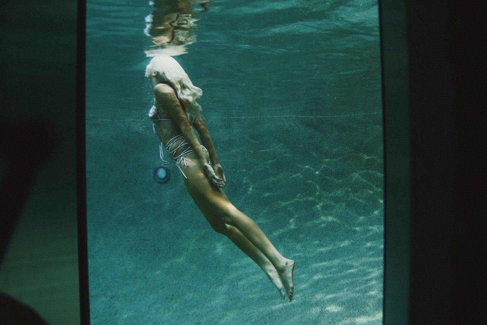 A model in a floral bikini diving underwater in a pool, seen through a window