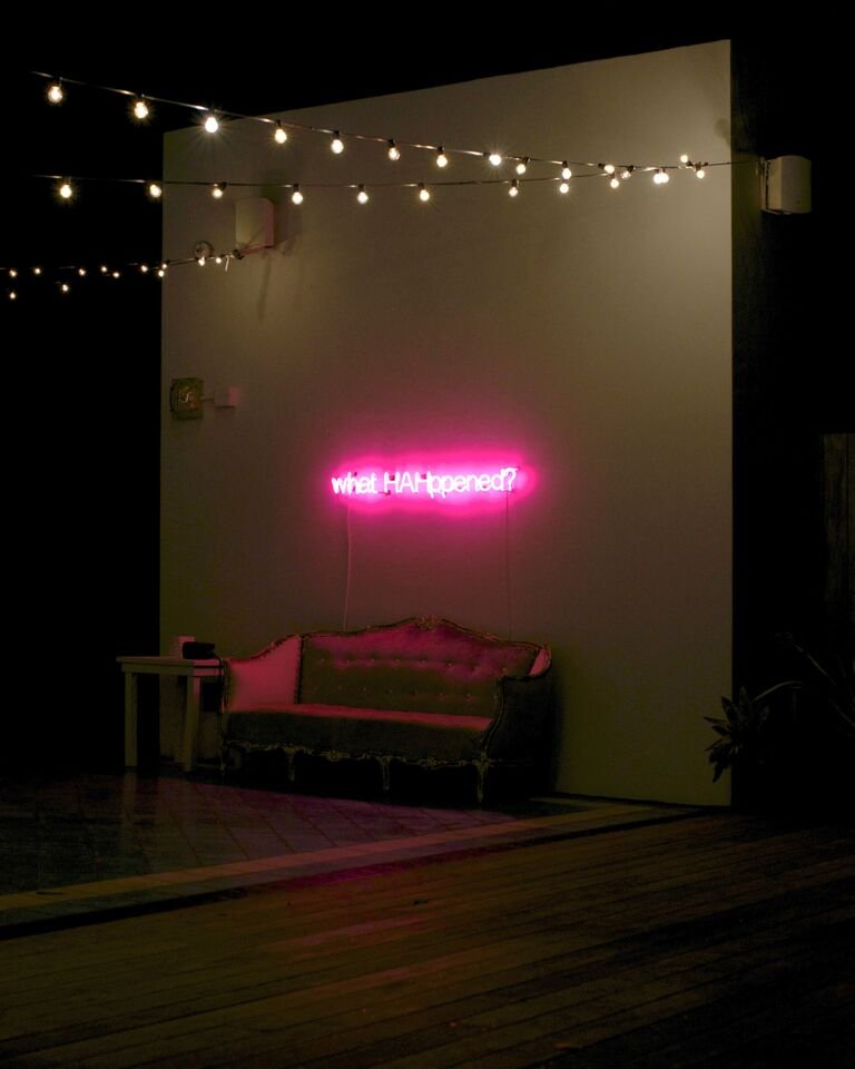 A pink neon sign reading &ldquo;what HAHppened?&rdquo; above a vintage tufted sofa on the rooftop at night, under string lights