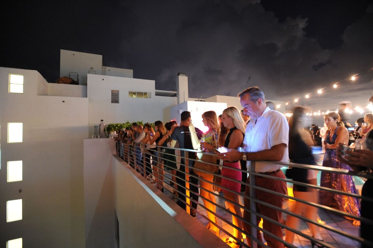 Guests lining a metal railing on the rooftop at night, looking out under string lights