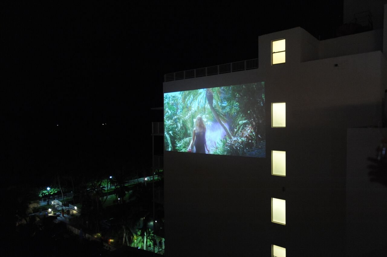 The brand film projected onto the side of a neighboring building at night, showing a woman among palm trees