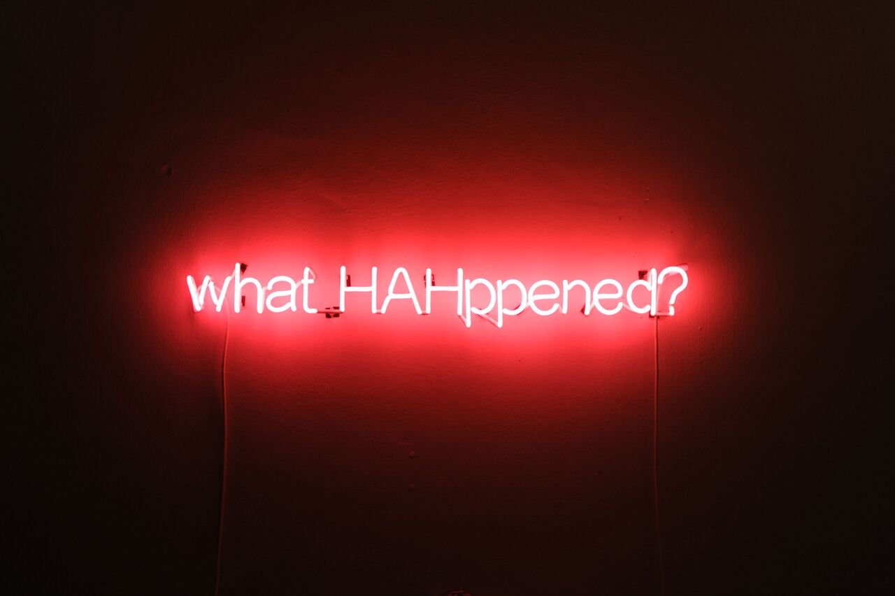 A red neon sign reading “what HAHppened?” on a dark wall