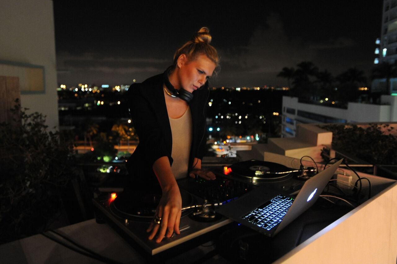 A DJ at turntables with a laptop on the rooftop, with city lights behind her