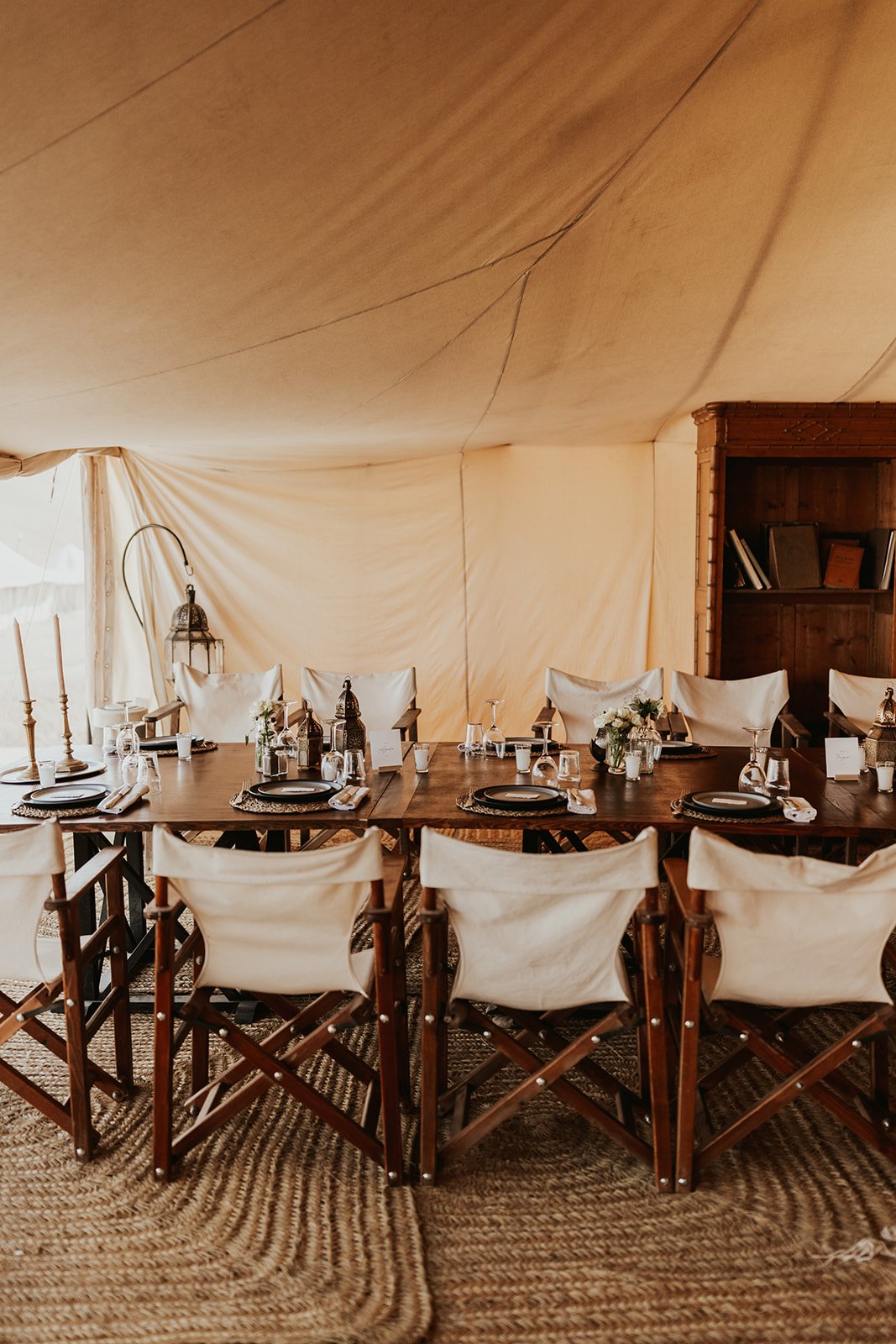 A long wood table set with black plates, brass lanterns, and white flowers beneath a cream canvas tent, with canvas director's chairs and a carved wood bookcase