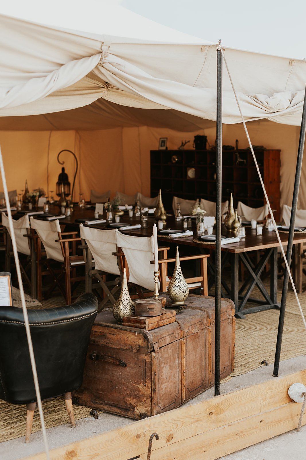 Brass vases and a candlestick on a worn leather trunk beside a black leather chair, in front of a dining table set under a canvas tent