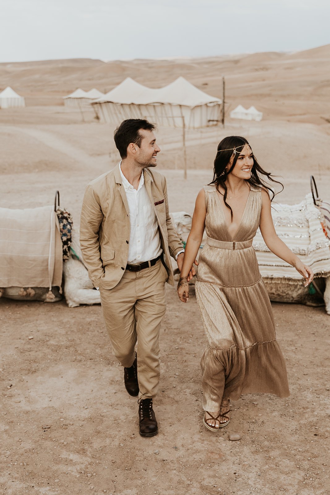 The couple walking hand in hand in the desert, with canvas tents and camel seats behind them