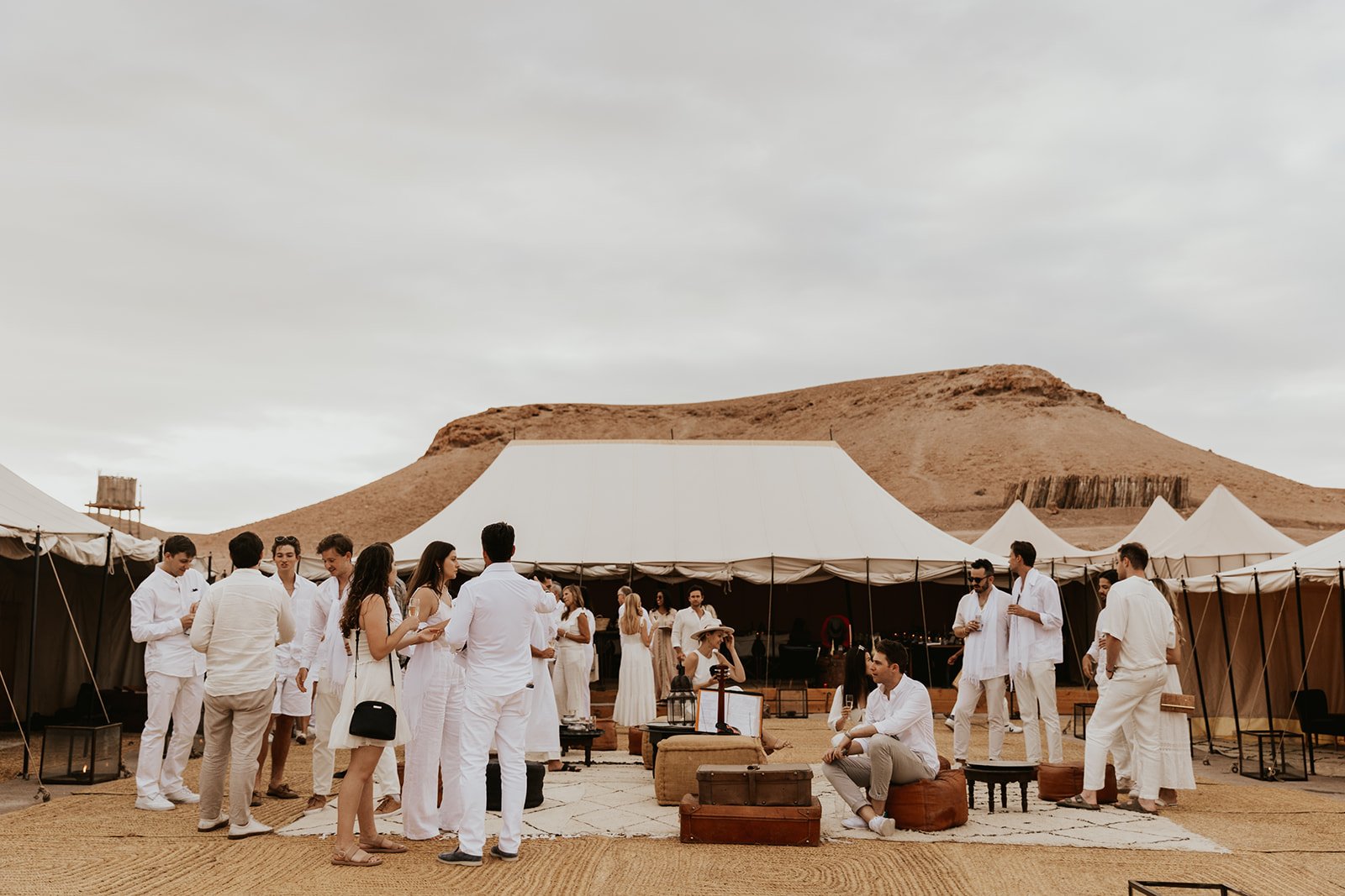 Guests dressed in white gathered on a rug with leather poufs and suitcases in front of a large canvas tent