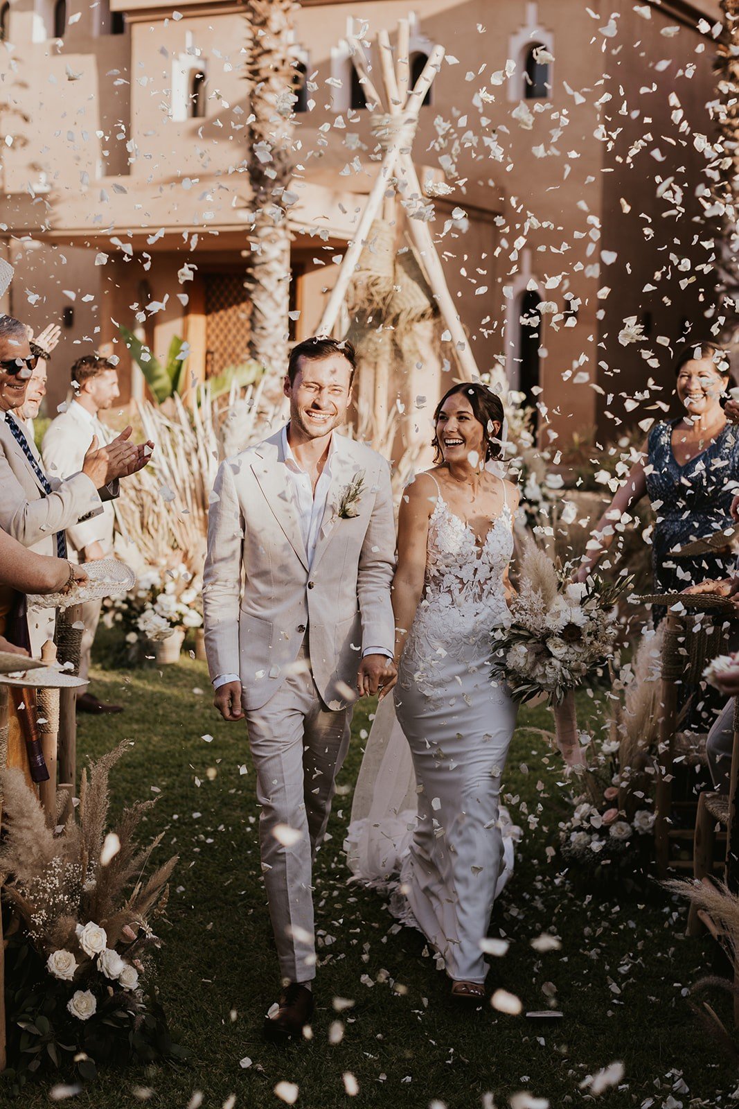 The couple walking up the aisle laughing as guests throw white petals, in front of a teepee frame and the villa