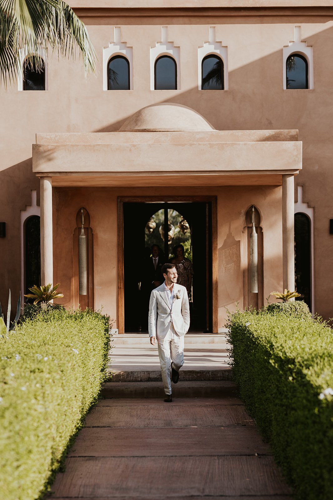 The groom in a light linen suit walking down a stone path between hedges from the villa entrance