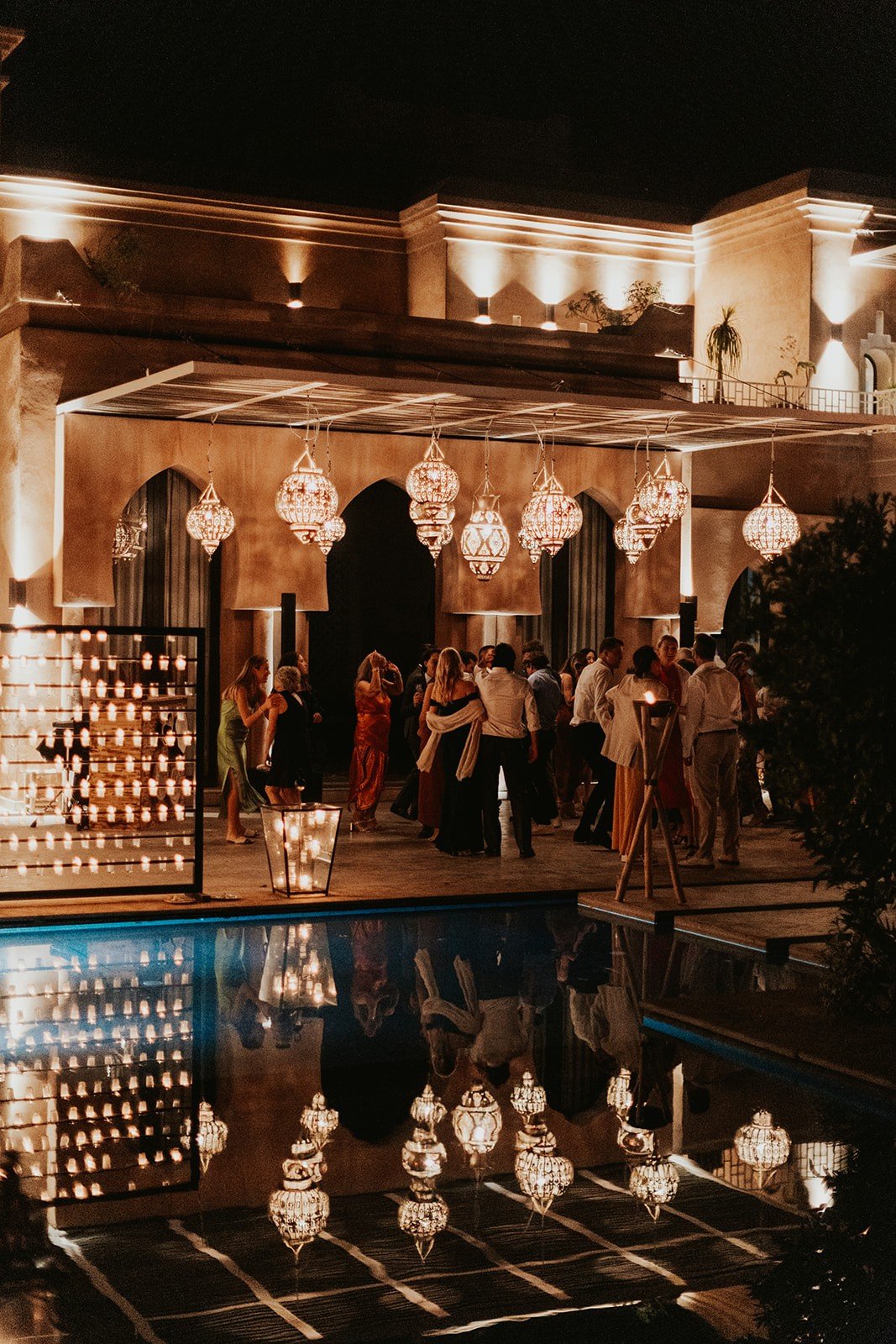 The villa at night with hanging brass lanterns, a wall of candles, and guests gathered beside the reflecting pool