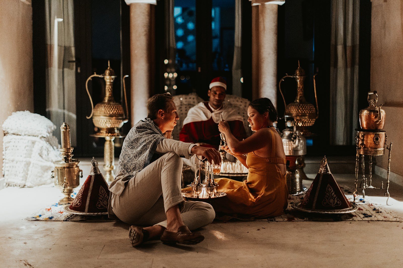 The couple sitting on a rug pouring Moroccan mint tea from a silver tray, with brass pitchers and a servant in a red cape behind them