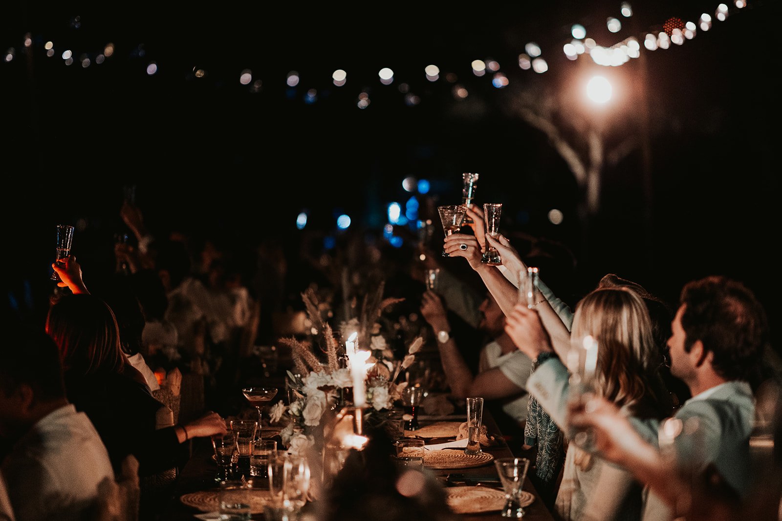 Guests raising glasses in a toast along a candlelit long table under string lights at night
