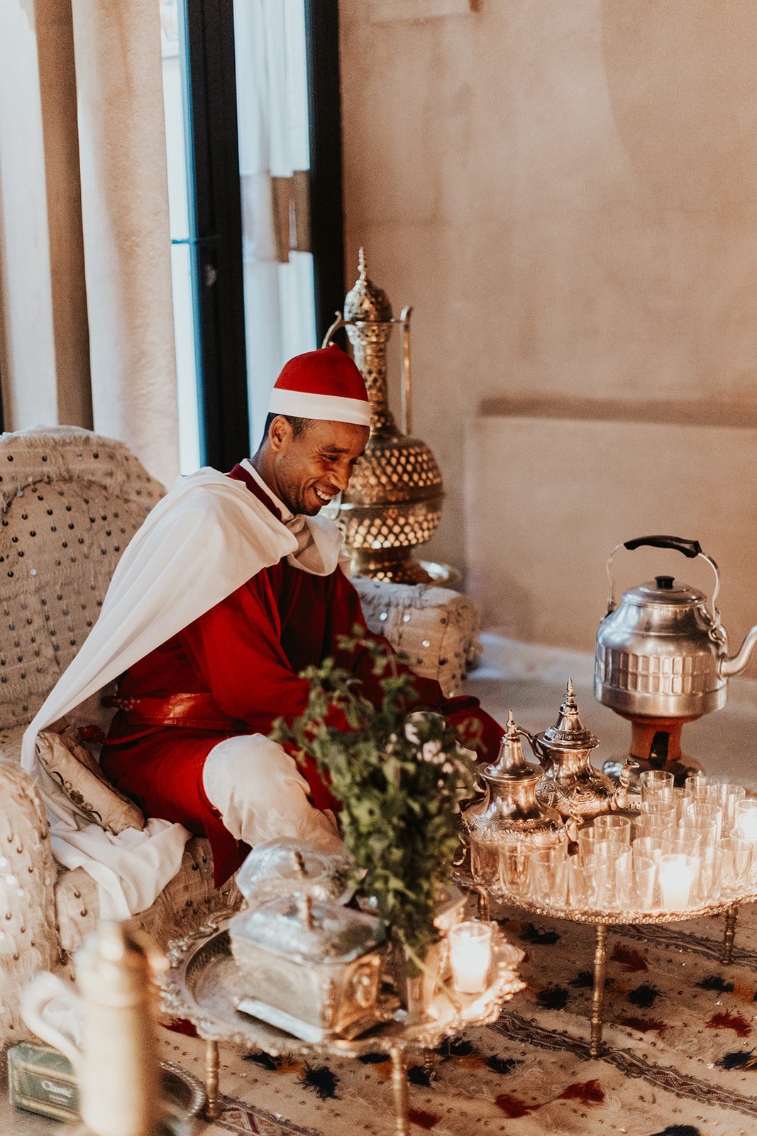 A man in a red cape and cap smiling beside a silver tea service, tall brass pitchers, and a tray of glasses on a patterned rug