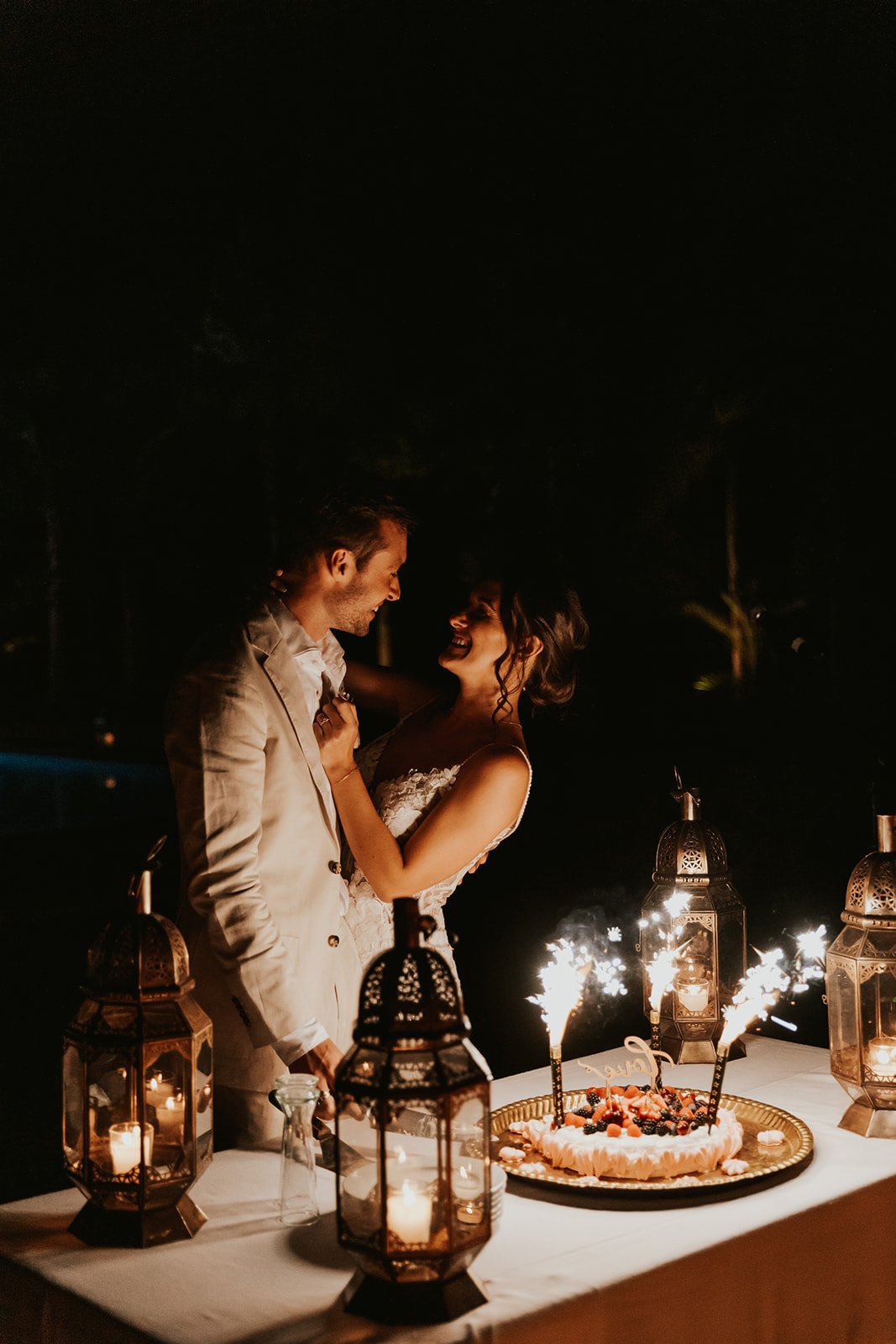 The couple embracing beside a cake topped with sparklers and berries, surrounded by brass lanterns at night