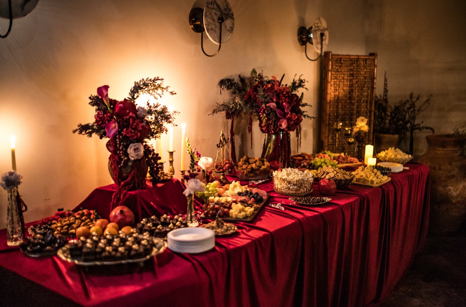 A buffet table draped in red velvet with candles, dark floral arrangements, pomegranates, cheeses, and sweets