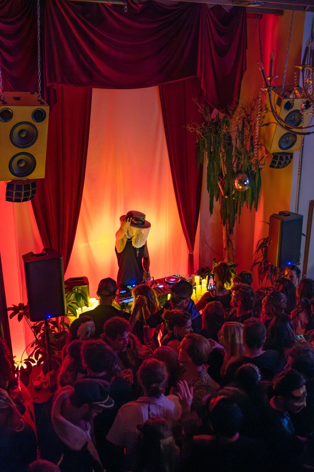 A DJ in a fringed hat playing for a packed crowd in front of red velvet curtains, hanging speakers, and a disco ball, seen from above