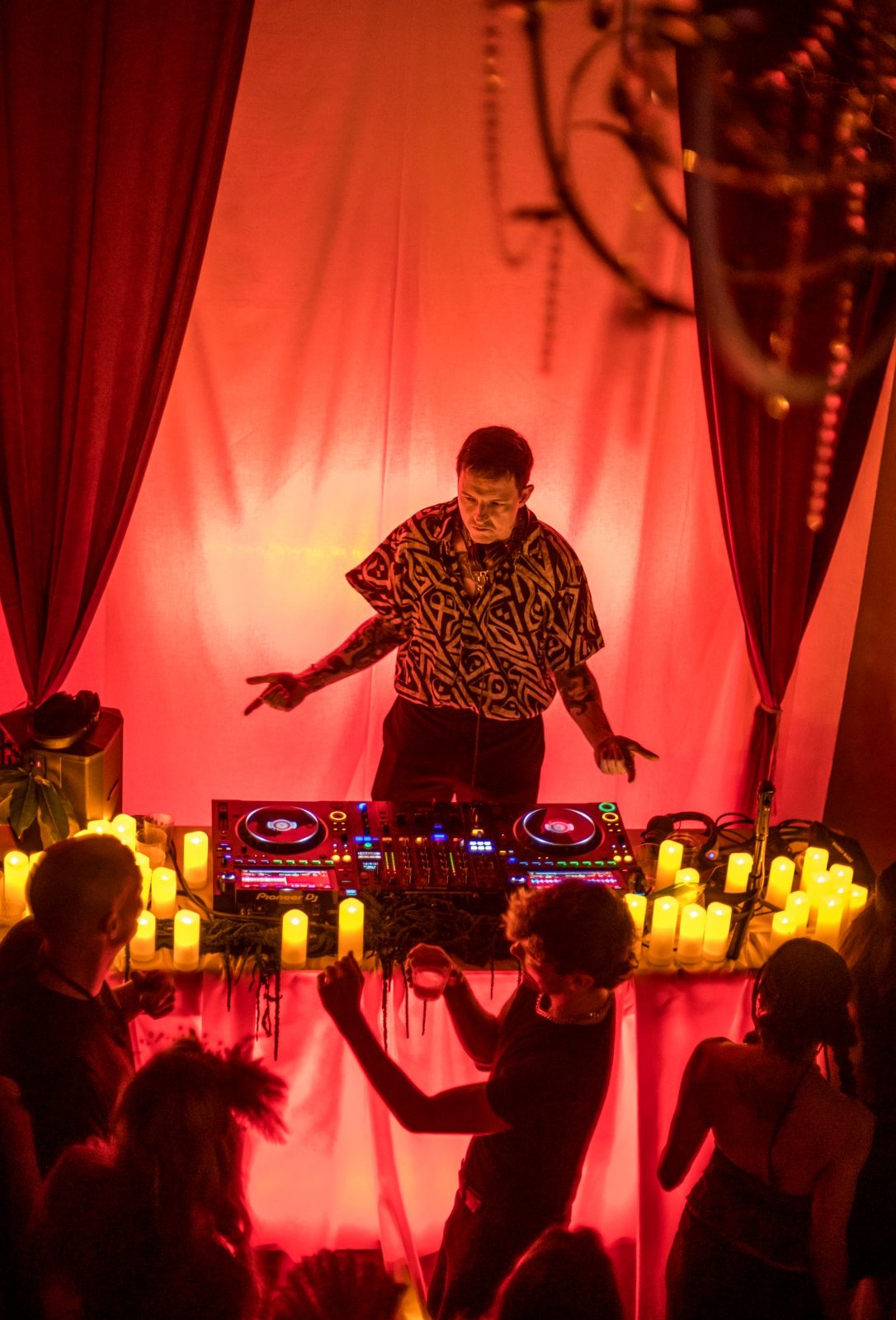 A DJ in a patterned shirt pointing at the crowd behind a table of turntables lined with candles