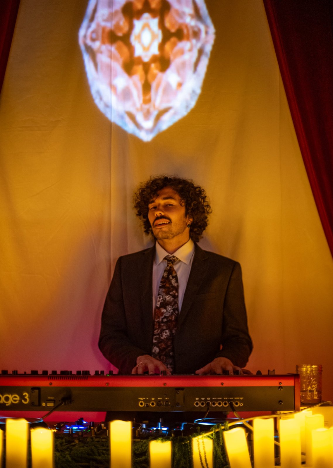 A keyboard player in a suit and floral tie in front of a projected kaleidoscope pattern