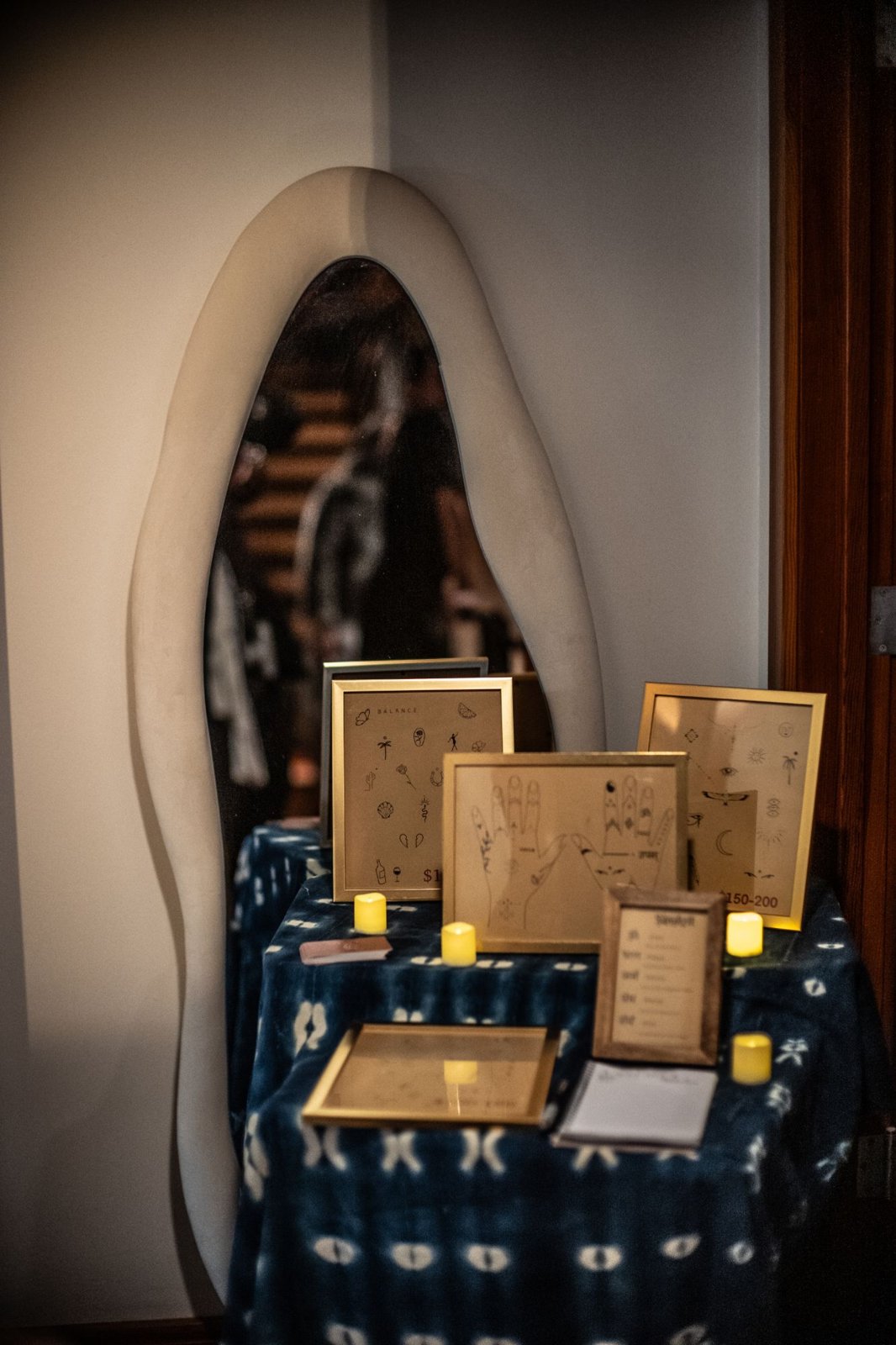 A table draped in indigo cloth with framed tattoo flash sheets and candles beside a sculptural mirror