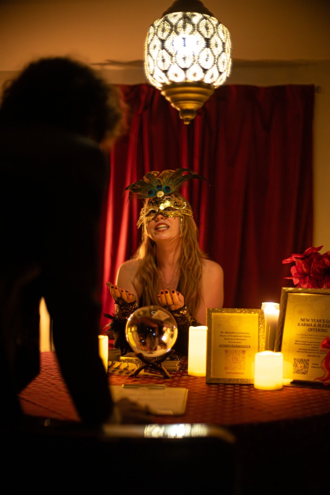 A fortune teller in a feathered mask with a crystal ball, tarot cards, and candles at a table